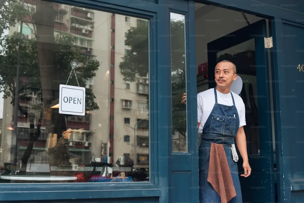 Chủ cửa hàng đứng trước cửa tiệm với biển Open